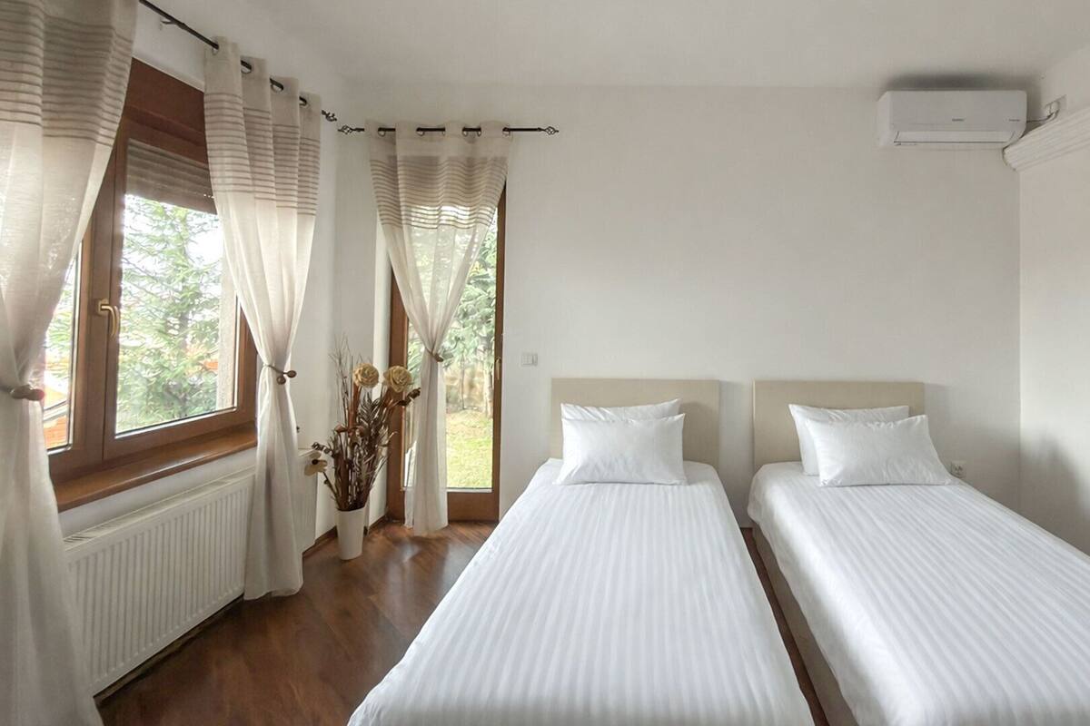 A hotel room with two single beds, a radiator, a window with curtains, and an air conditioning unit.