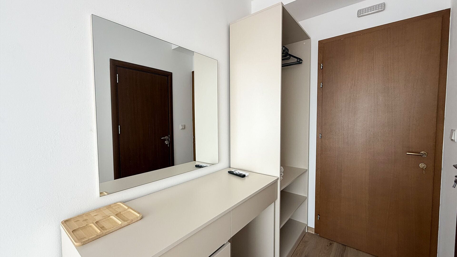 A room with a white console table, a mirror, a wooden door, and a shelf with a remote control.
