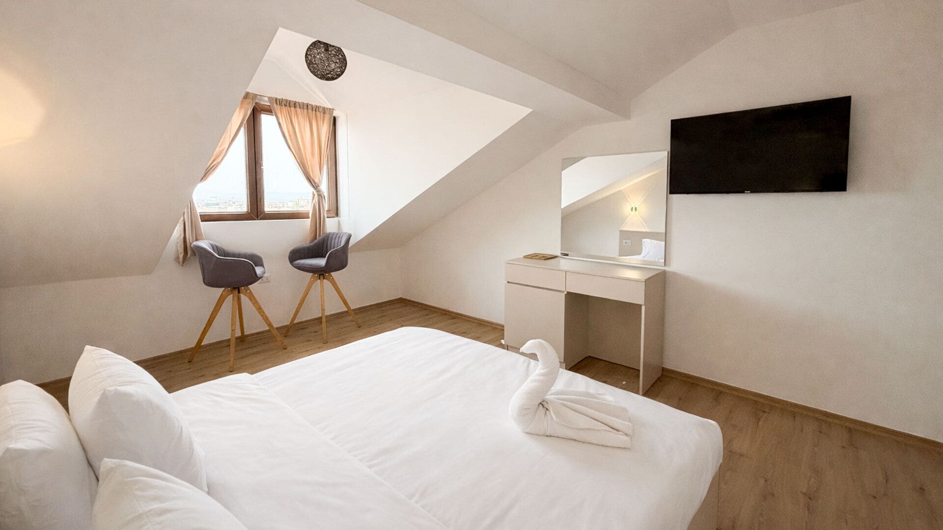 A modern hotel room with a large bed, a desk, two chairs, a TV, and a window with curtains.