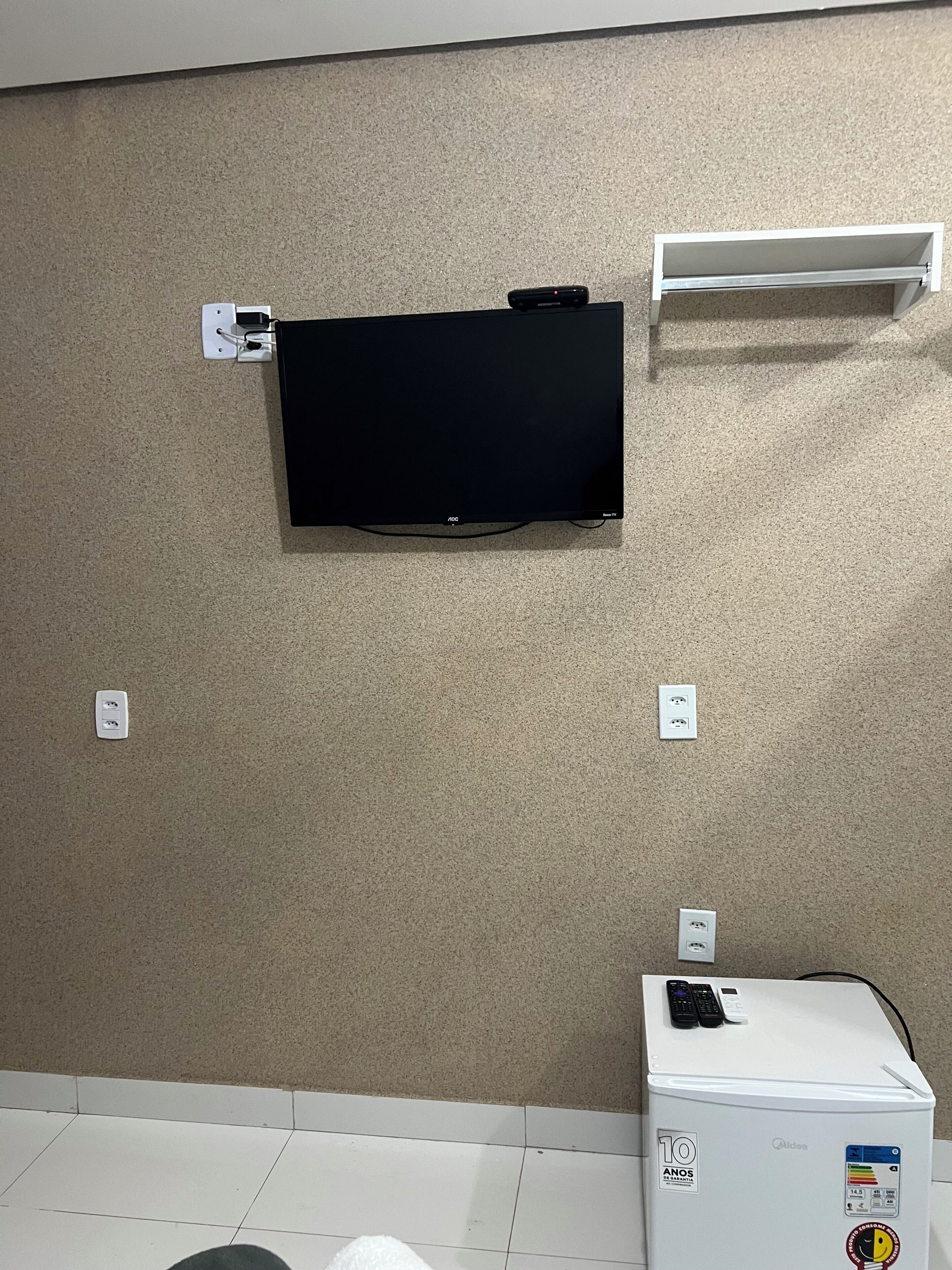A room with a flat-screen TV mounted on the wall, a white refrigerator, and a white shelf unit.