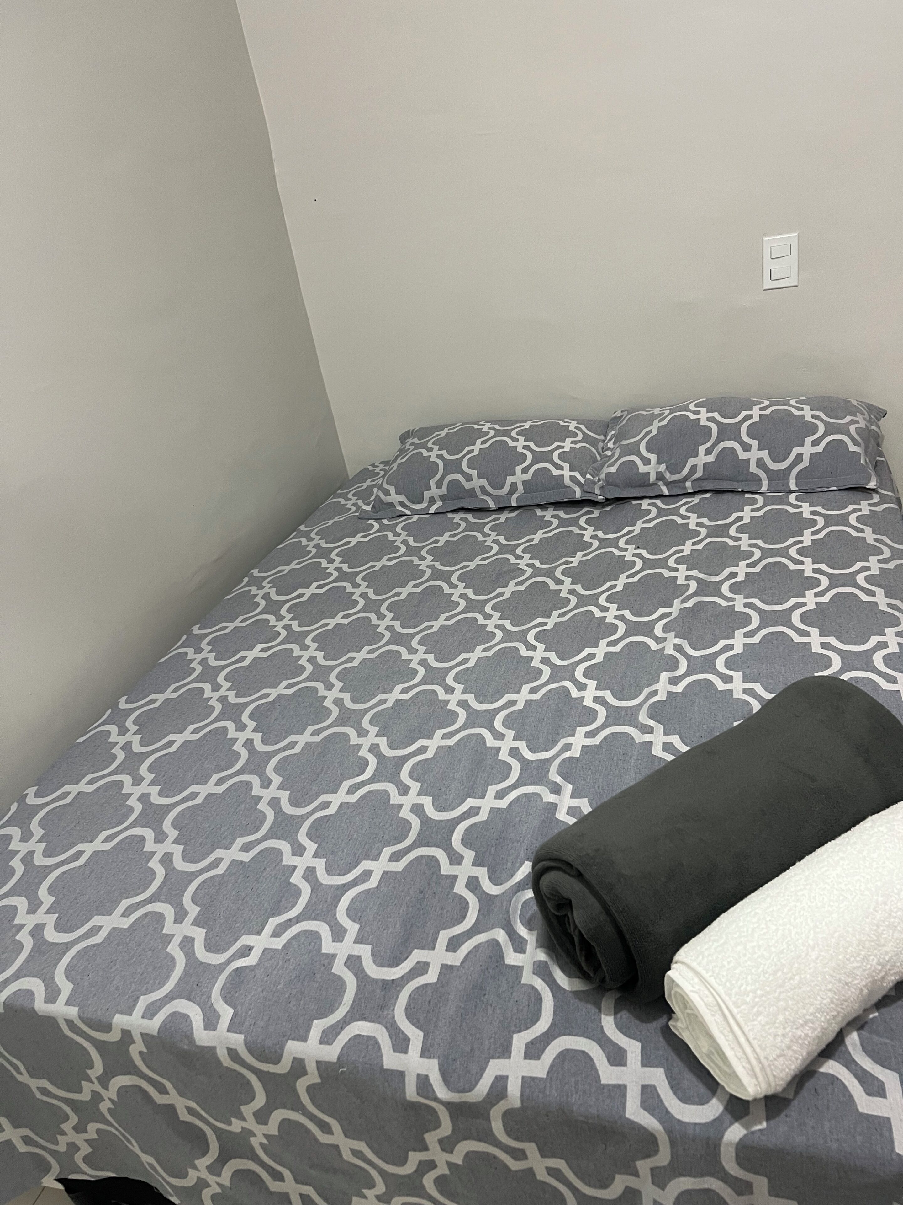 A single bed with a patterned bedspread, a folded dark towel, and a rolled white towel.