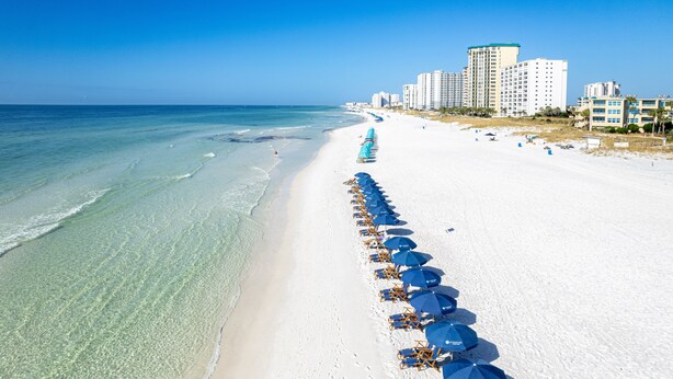 Une plage bordée de rangées de parasols bleus et de chaises longues, un ciel bleu dégagé et des bâtiments en arrière-plan.