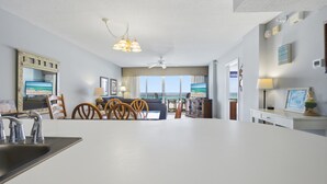 Una sala de estar moderna con una gran mesa blanca, un ventilador de techo y vistas al océano a través de amplios ventanales.