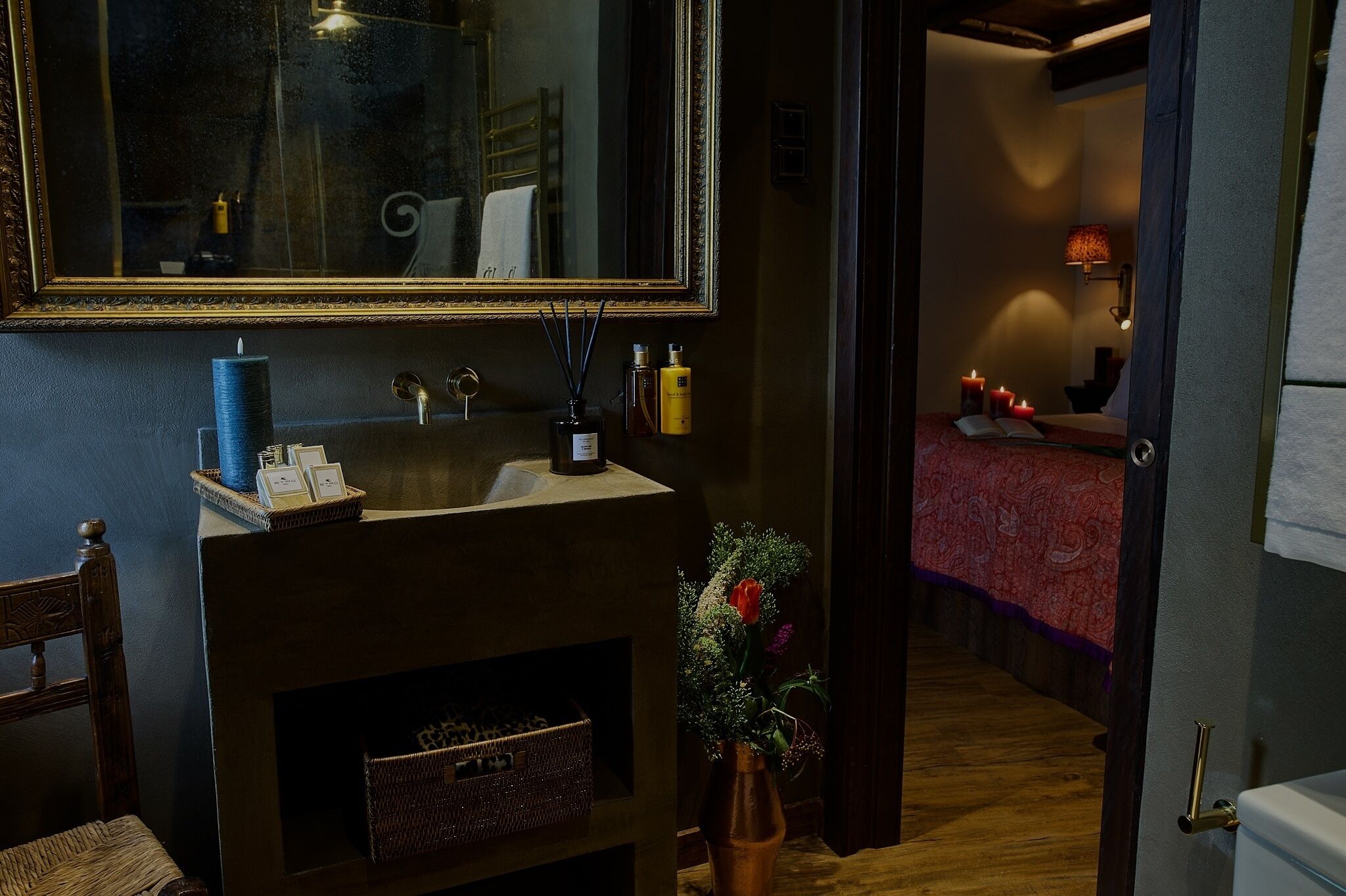 Une salle de bain avec un grand miroir, un lavabo, un vase de fleurs et une chaise en bois.