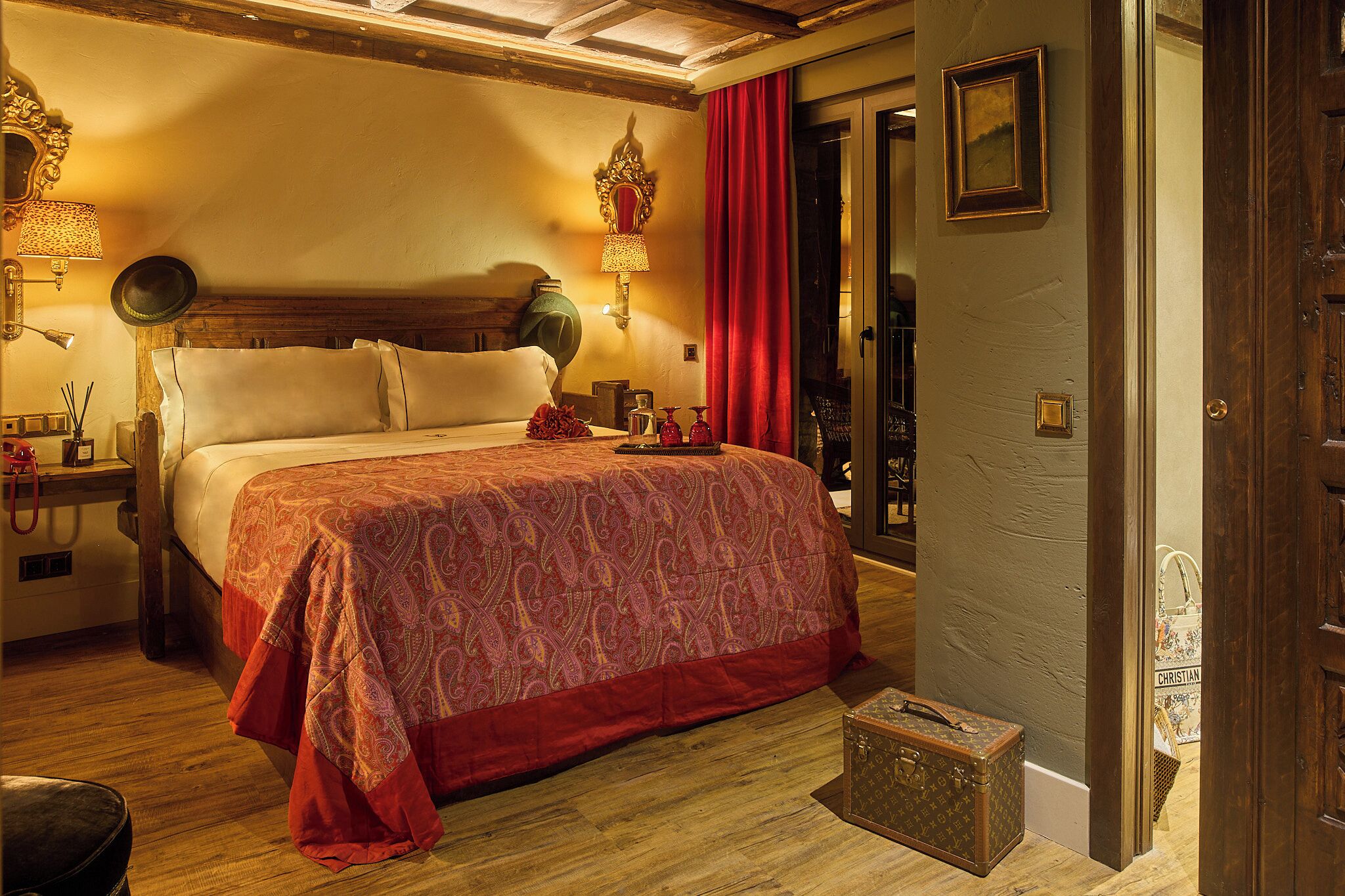 A hotel room with a large bed, a wooden headboard, a nightstand with a lamp, and a red curtain.