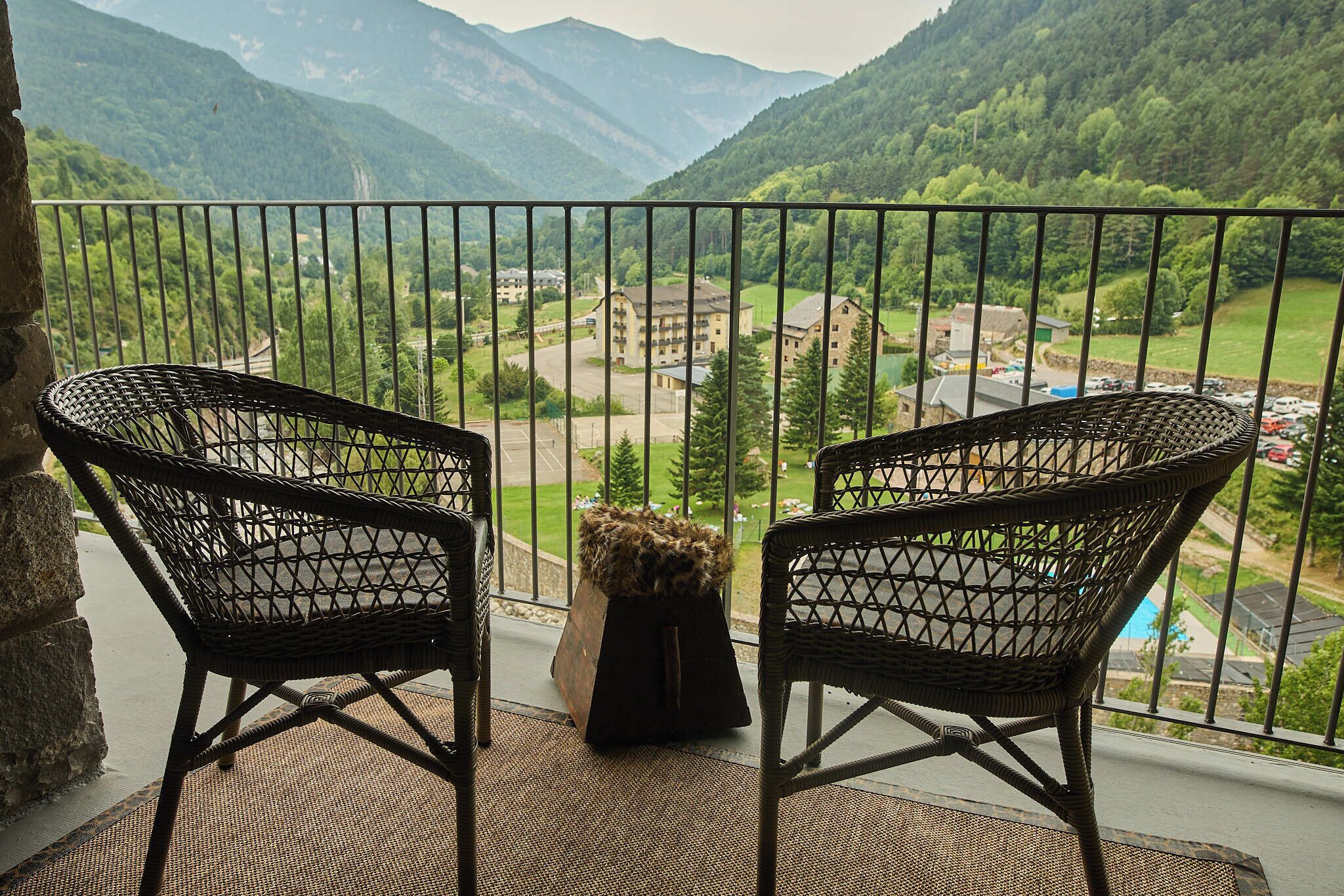 A balcony with two wicker chairs, a small table with a plant, and a view of a valley with buildings and a road.