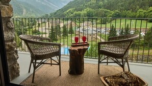 A balcony with wicker chairs, a small wooden table, and a view of a valley with buildings and a river. - Hotel Valle de Pineta (Bielsa)