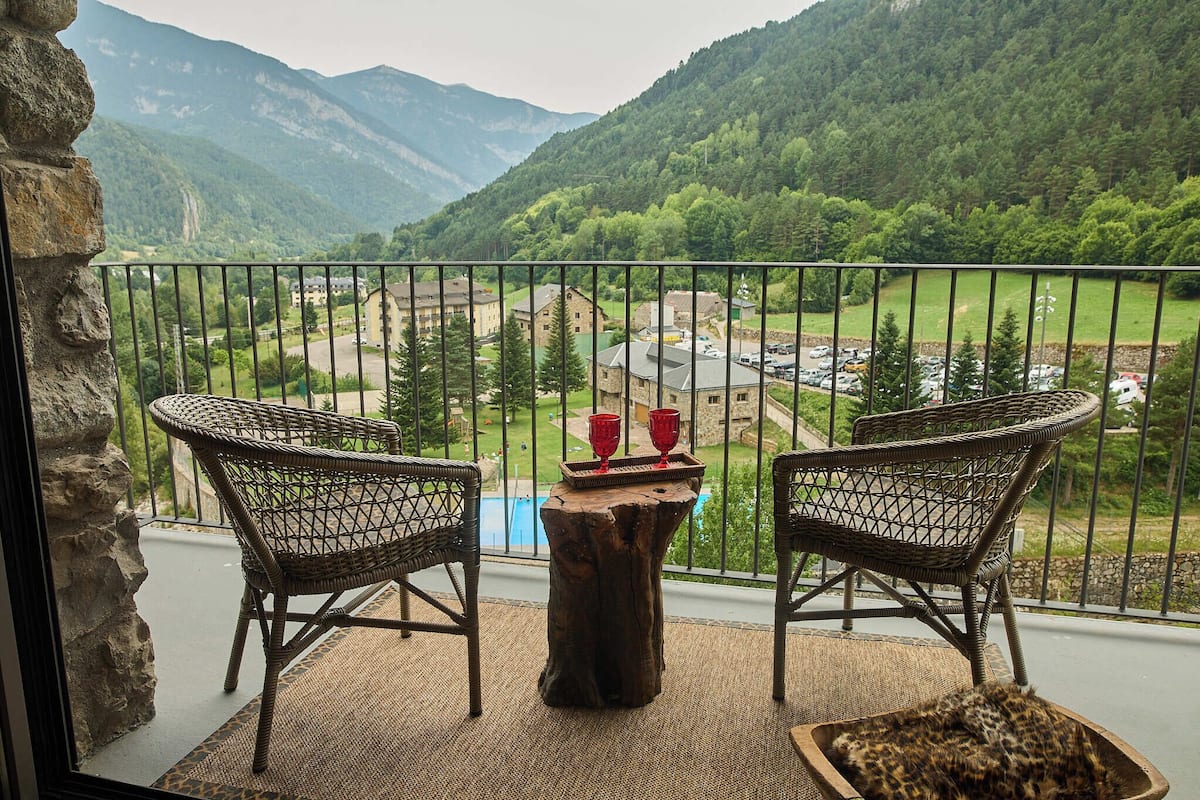 Un balcón con sillas de mimbre, una mesa de madera y vistas a un valle con montañas y un estacionamiento.