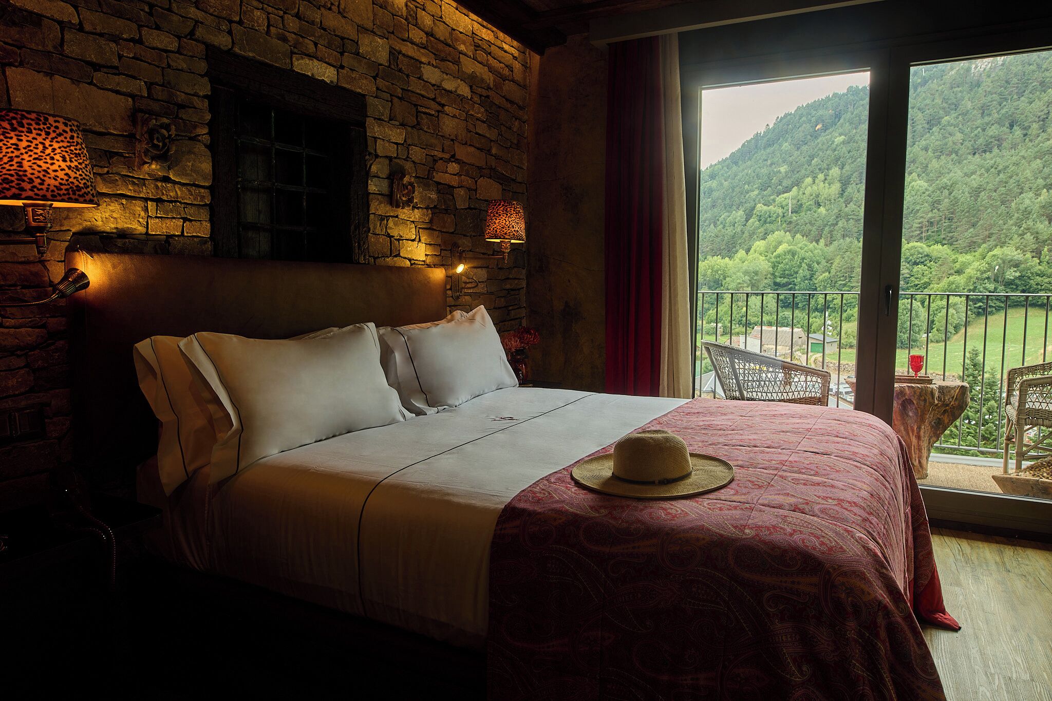 A bed with a patterned bedspread, a hat, and a view of a balcony with chairs.