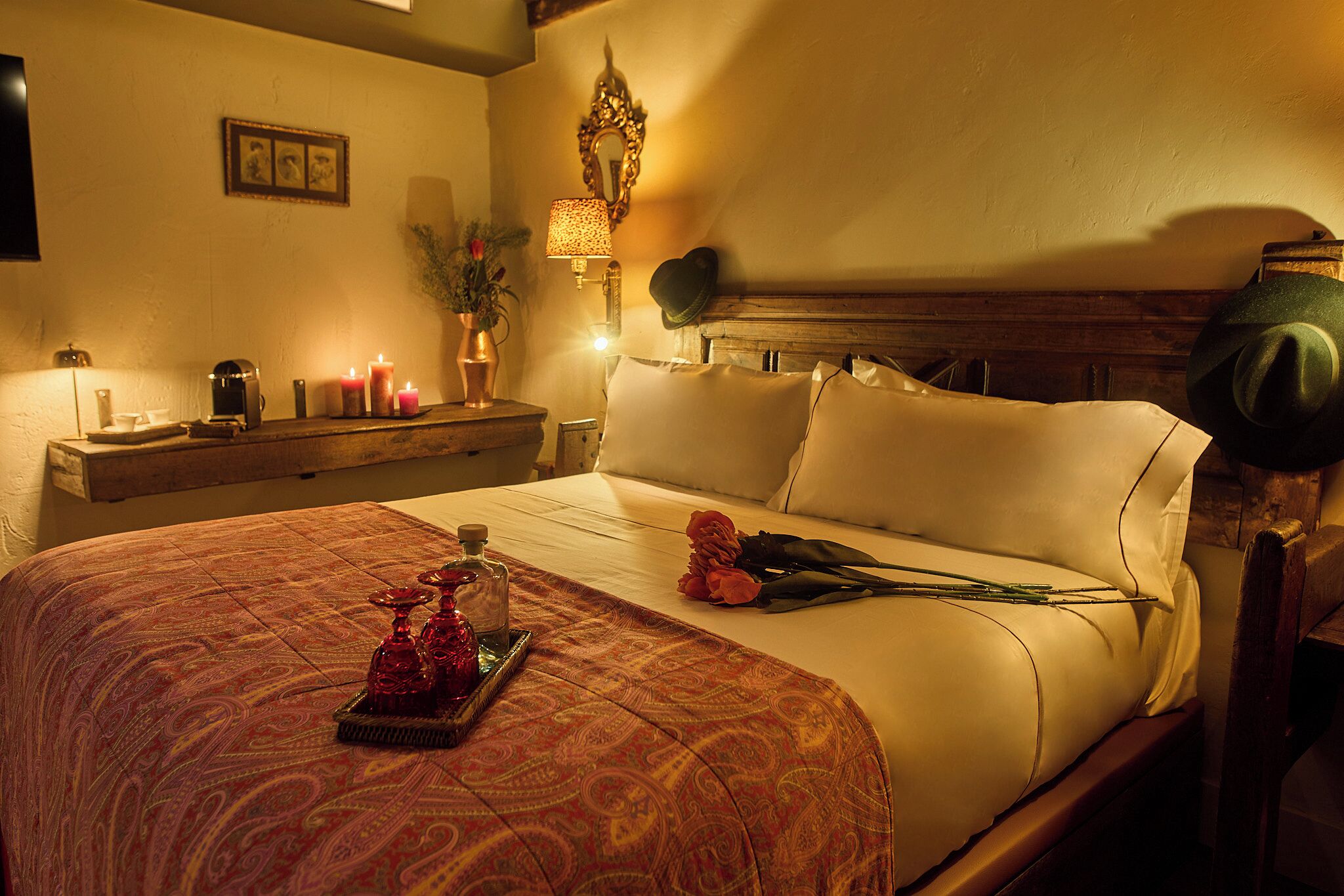 Una cama bien hecha con una colcha estampada, una mesita de noche con lámpara, velas y un jarrón con flores.