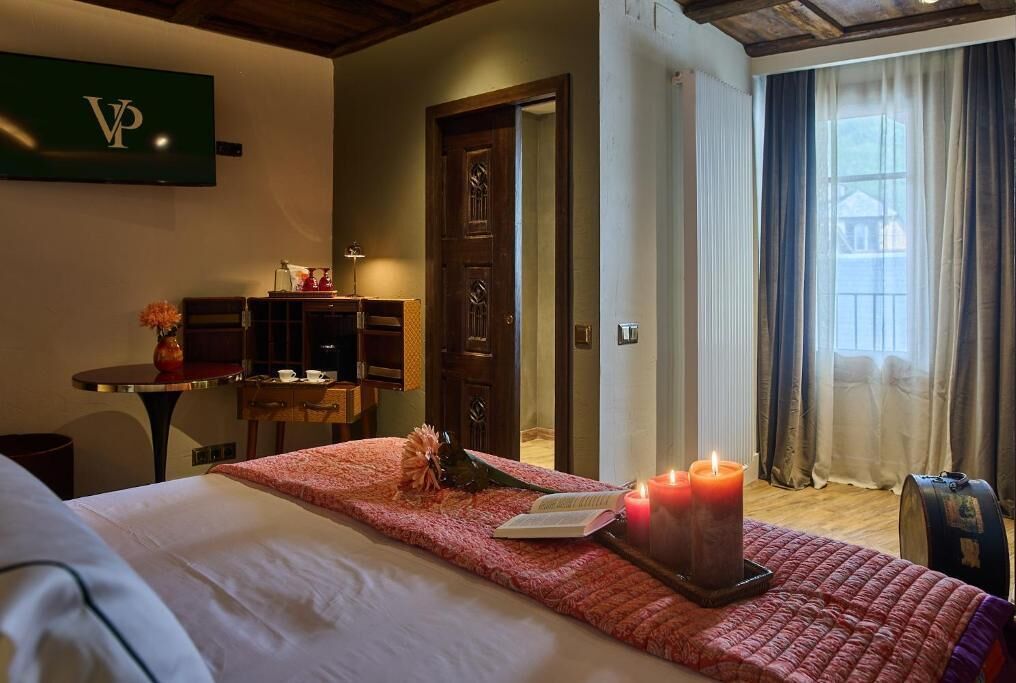 Hotelkamer met een bed, nachtkastje met een boek en een kaars, bureau met een lamp, en uitzicht buiten door een raam.