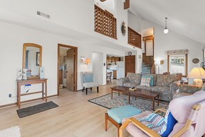 A spacious living area with a high ceiling, wooden furniture, a kitchen in the background, and a view of the outside. - Just Beachy: Short walk to Beach | Hot Tub (Waves)
