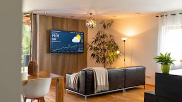 A modern living room with a wooden floor, a black sofa, a white chair, a dining table, a wall-mounted TV displaying a graph, and a potted plant.