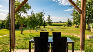 Eine Außenpatio mit Tisch und Stühlen, umgeben von einer Holzpergola, mit Blick auf eine Rasenfläche mit Bäumen und eine ferne Feldlandschaft.