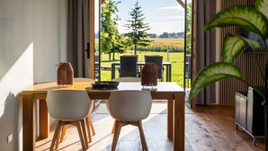 Ein Essbereich mit Tisch und Stühlen, Blick auf eine grüne Landschaft durch eine offene Tür und ein Blumentopf.