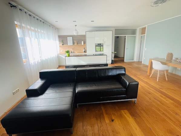 Un salon moderne avec un canapé en cuir noir, un parquet en bois, un coin repas avec une table et des chaises blanches, et une cuisine en arrière-plan.
