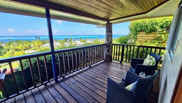 Eine Holzterrasse mit Geländer, Korbmöbeln und Blick auf das Meer und viel Grün.