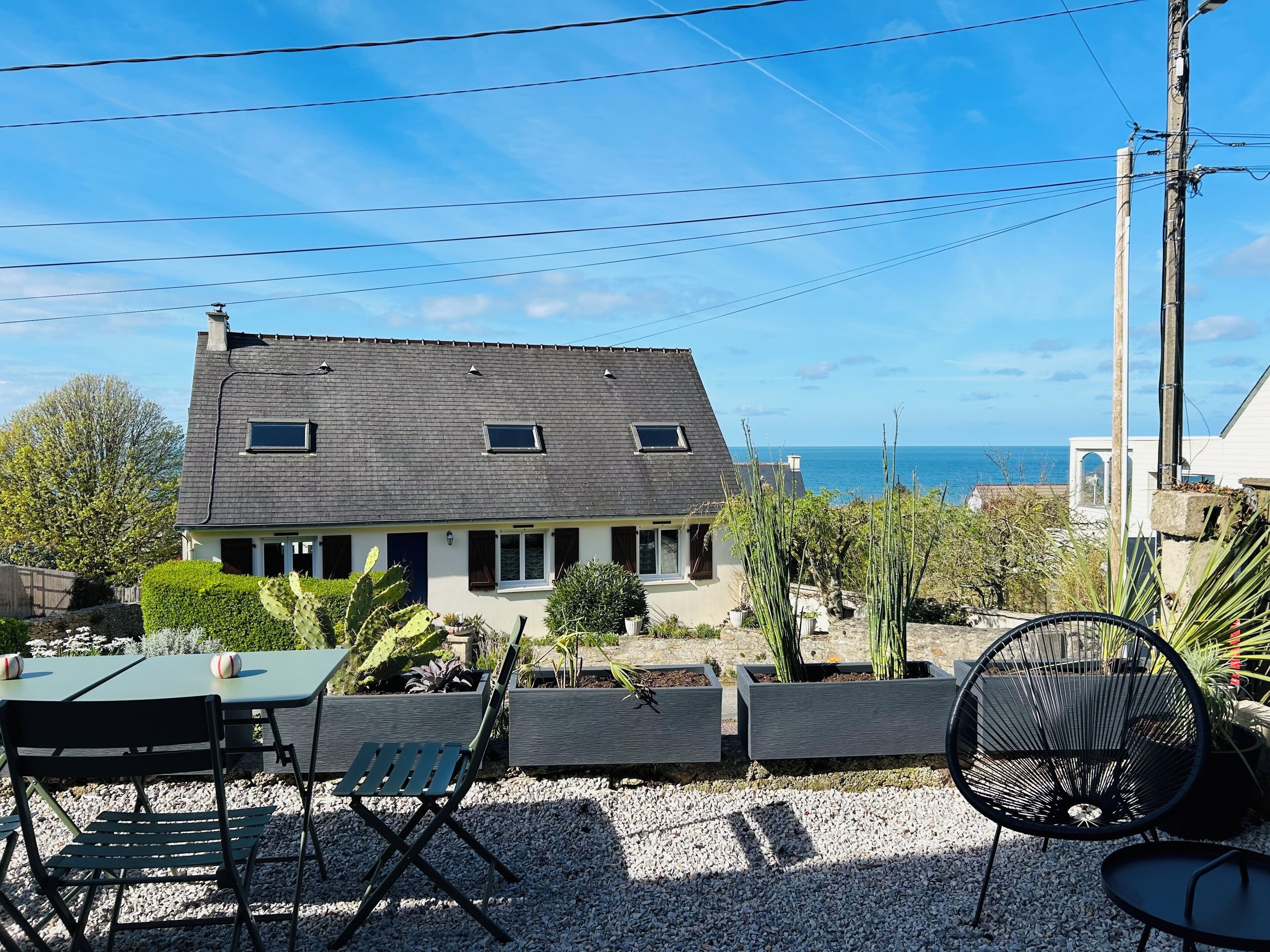 A house with a sloped roof, a patio with chairs and a table, and a view of the sea.