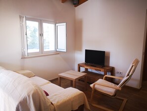 Una habitación con cama, televisión sobre un soporte de madera, una silla y una ventana con contraventanas abiertas.