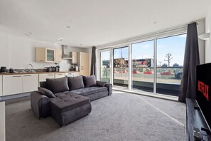 A modern living room with a grey sofa, a kitchen area, and a large window with a view of the outside. - ExCel & O2 Waterfront Apartment with Gym Access (Greater London)