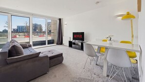 Una sala moderna con un sofá gris, una mesa de comedor con sillas amarillas, un televisor mostrando 'NETFLIX' y grandes ventanales con vistas a la ciudad.