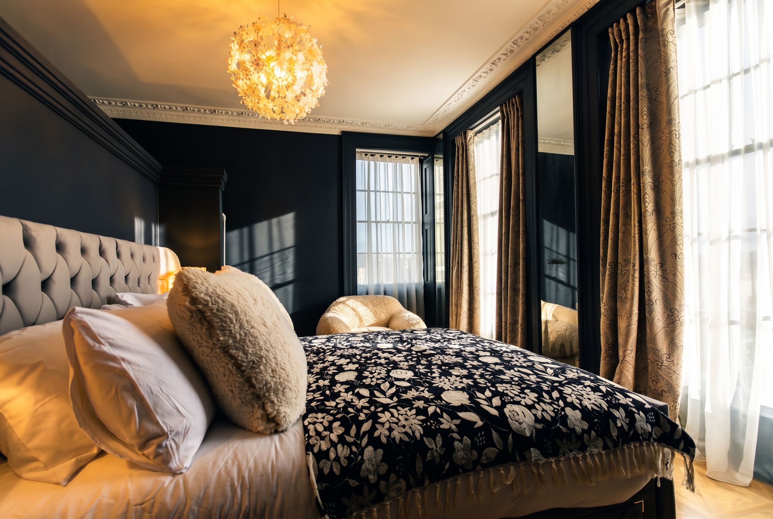 A bedroom with a tufted headboard, a large bed with floral bedding, a hanging light fixture, and a view through a window with sheer curtains.