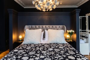 Ein Schlafzimmer mit einer gebügelten Kopfteil, einer zudeckenden Bettdecke mit Blumenmuster, zwei Nachttischlampen und einer Kristallleuchter.