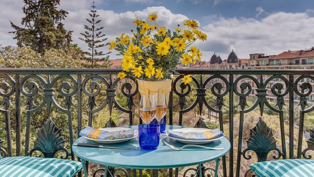 Een tafel met een vaas gele bloemen, twee wijnglazen en borden op een balkon met een sierhek van ijzer en uitzicht op gebouwen.