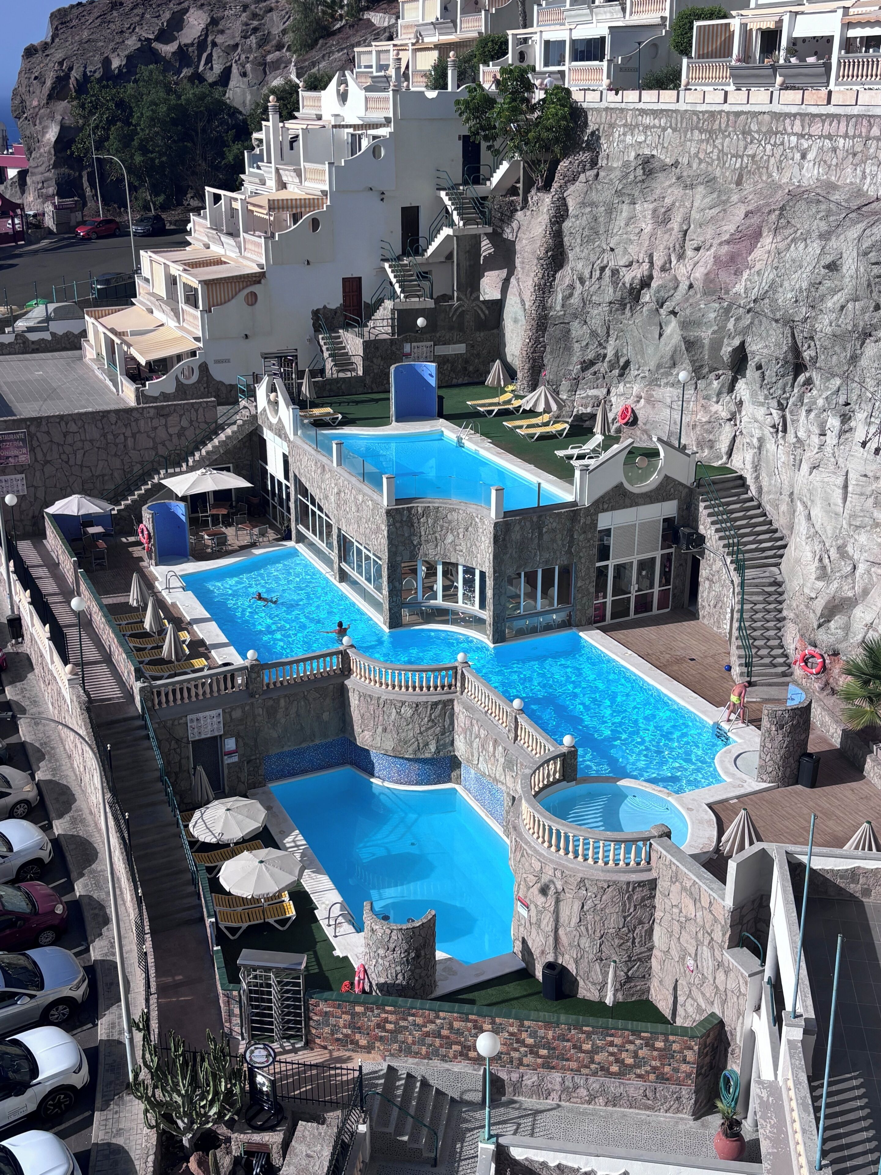 A multi-level swimming pool complex with stone architecture, multiple levels of balconies, and a view of the surrounding rocky landscape.