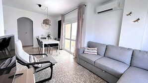 En moderne stue med en grå sofa, et spisebord med stole, et lille køkkenområde og et stort vindue med gardiner.