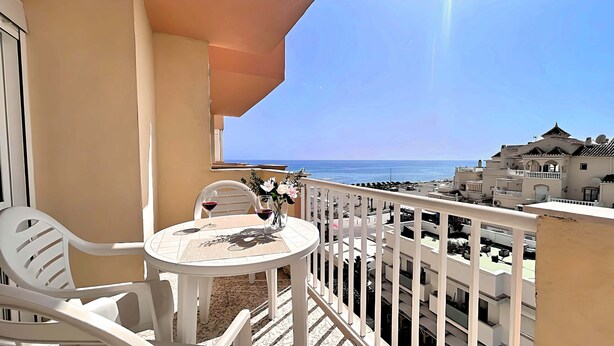 En balkon med et bord og stole, et glas rødvin og udsigt til havet og bygninger.
