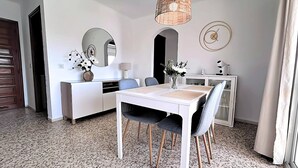 Et spiseområde med et hvidt bord, grå stole, en hvid kommode, et rundt spejl, et vægur og en vævet lampe.