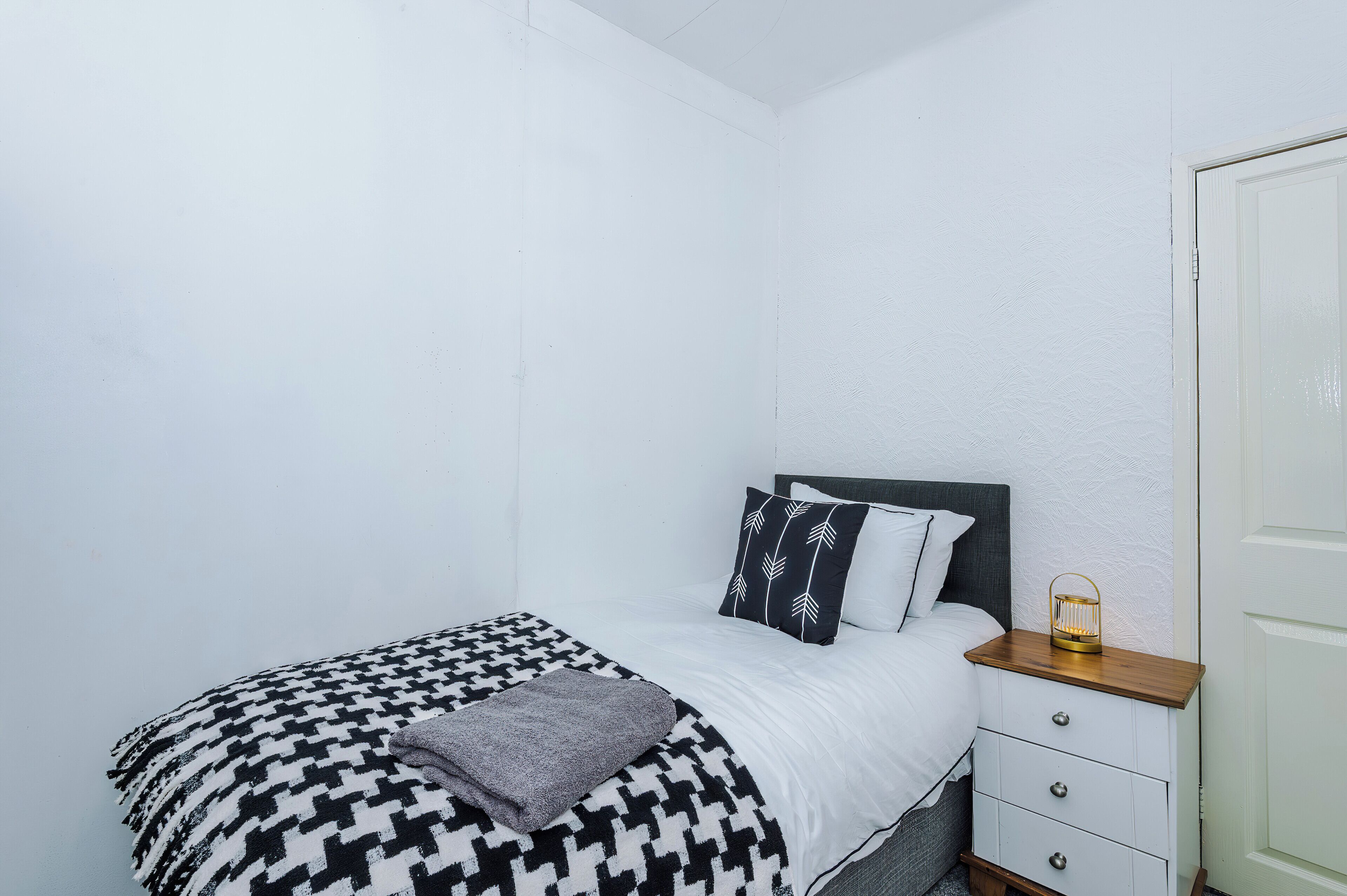 A single bed with a patterned bedspread, a bedside table with a lamp, and a door to the right.