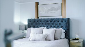 A neatly made bed with a tufted headboard, white bedding, and a bedside table with a lamp and a small decorative item. - Luxury 3 Bed - 6 min to Airport  (Cheadle)