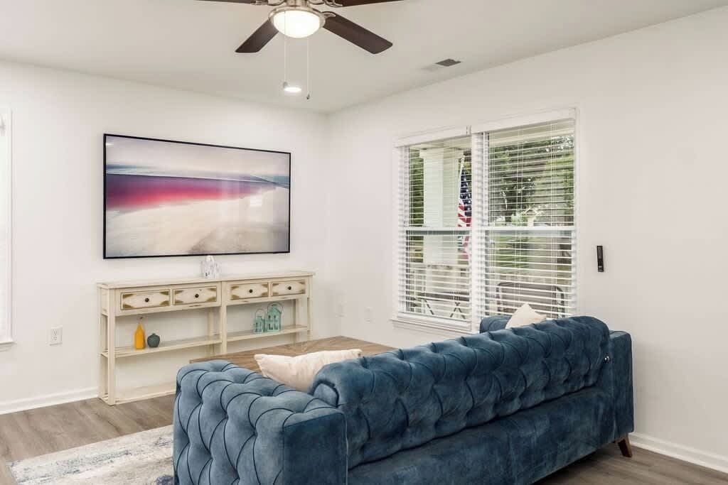 Un salon avec un canapé bleu, un téléviseur fixé au mur, une console blanche et une fenêtre avec des stores.