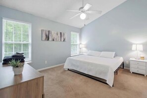 Une chambre à coucher comprenant un lit, des tables de chevet, un ventilateur de plafond et deux fenêtres équipées de stores.