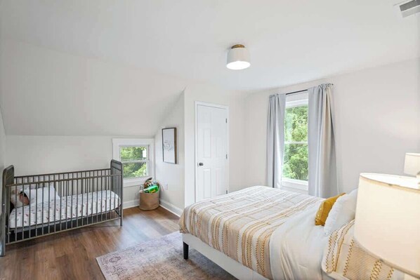 Ein Schlafzimmer mit einem Kinderbett, einem Bett, einem Fenster mit Vorhängen und einer Tür.