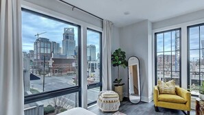 Habitació d'hotel moderna amb grans finestres, un llit, una butaca groga, un mirall i una planta en test.