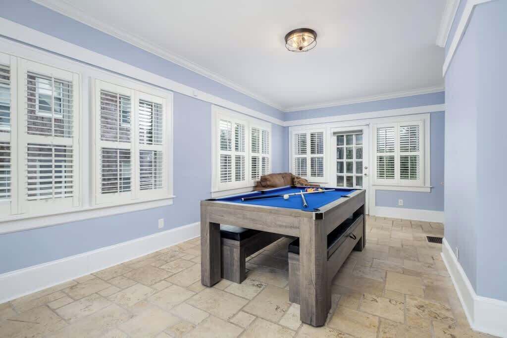 A pool table in a room with light blue walls and white trim, large windows with shutters, and a stone-tiled floor.