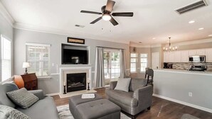 A modern living room with a fireplace, grey sofas, a ceiling fan, and a kitchen area with a microwave. - Pool is Open! 4BR Retreat • Gameroom • Sleeps 12 (Charlotte)