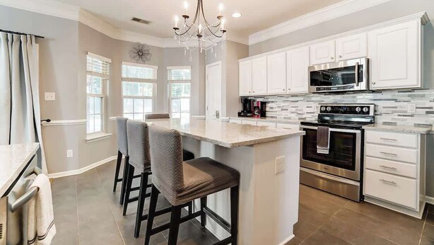 A kitchen with white cabinets, a central island with bar stools, a chandelier, and stainless steel appliances. - Pool is Open! 4BR Retreat • Gameroom • Sleeps 12 (Charlotte)