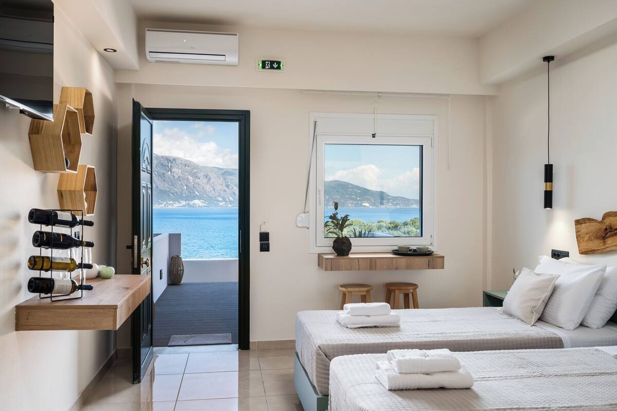 A hotel room with a bed, a window with a view of the sea, a shelf with wine bottles, and a wooden wall decor.