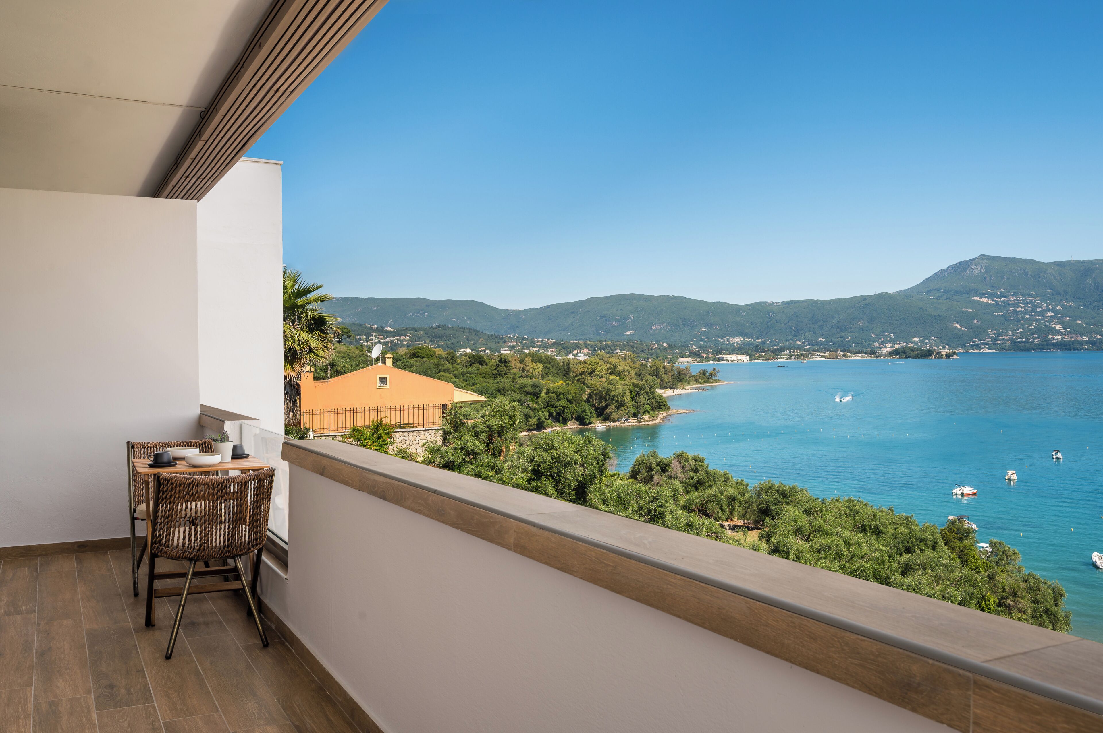 A balcony with a table and chairs, overlooking a coastal area with mountains and a clear sky.