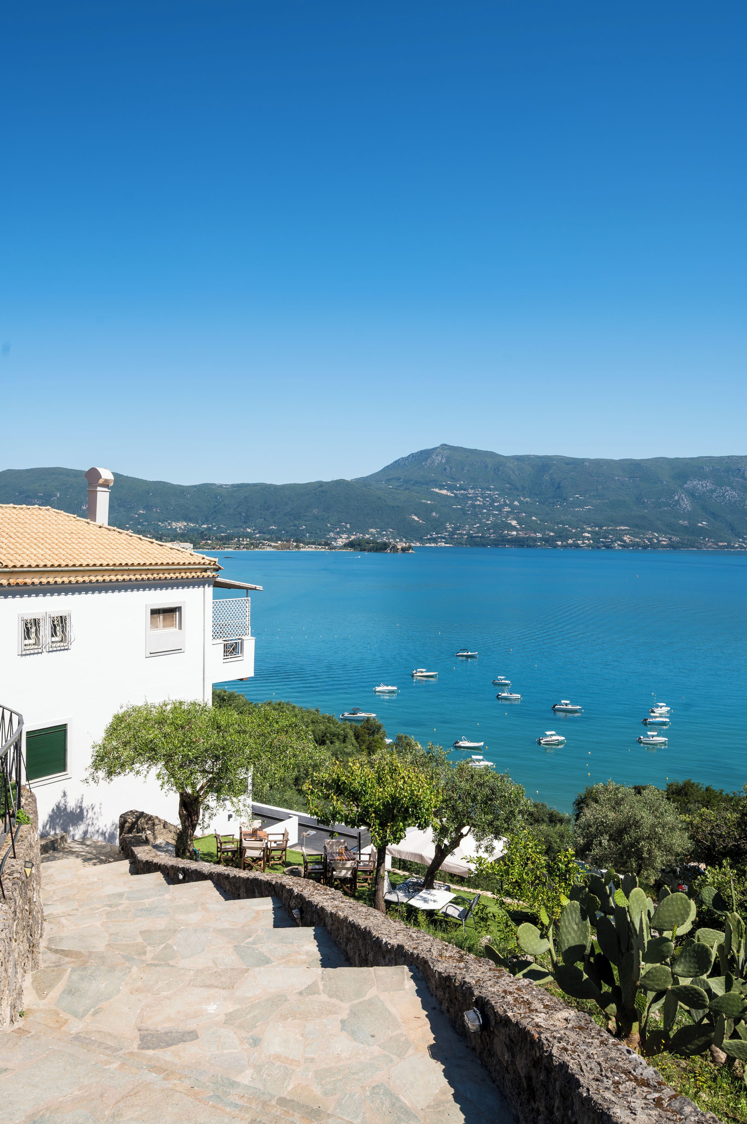 A coastal property with stone steps leading to a white building, a clear blue sea with boats, and a mountainous backdrop.