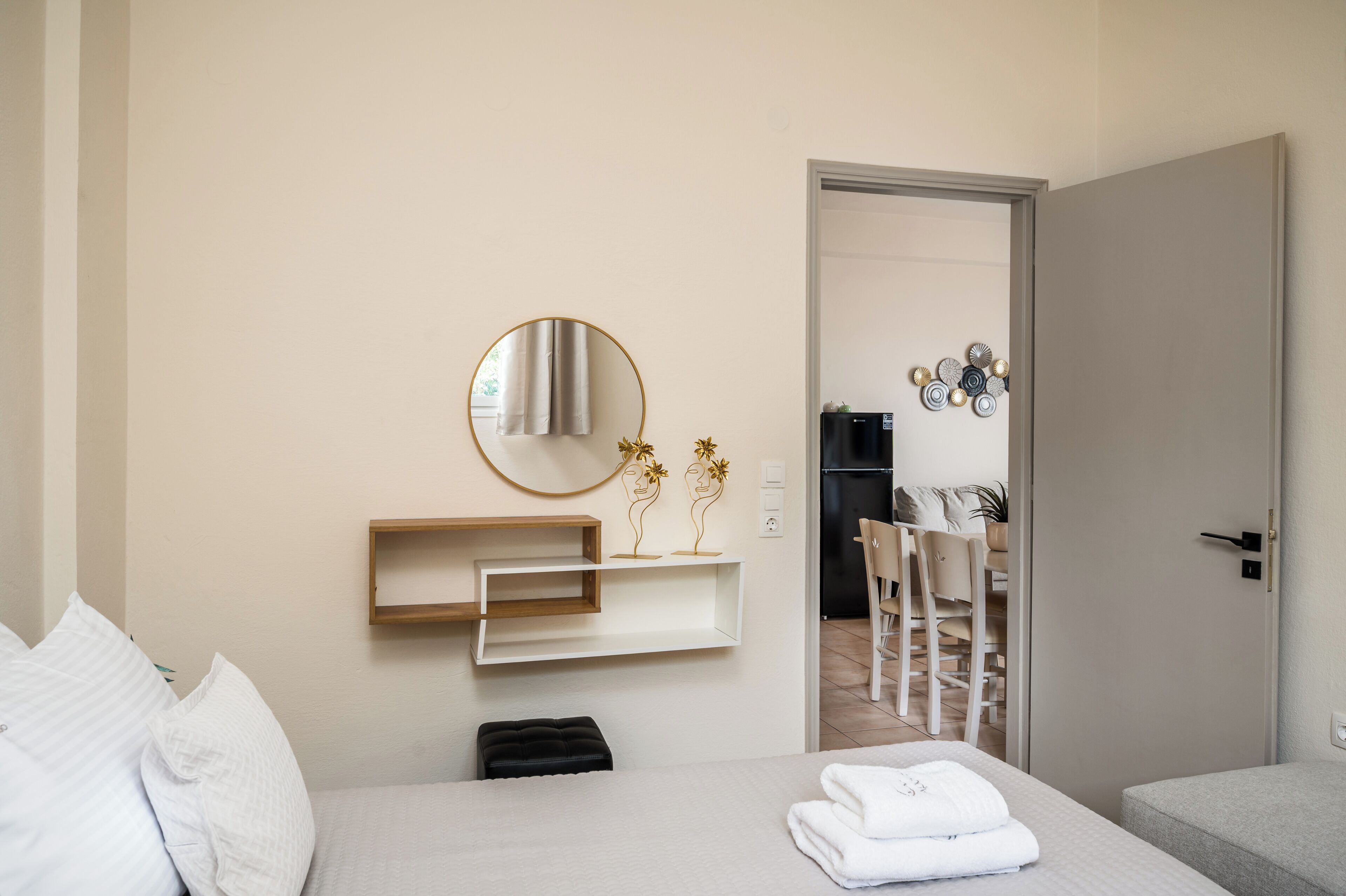 A modern hotel room with a bed, a bedside table with a lamp, a mirror, and a small shelf with decorative items.