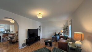 A modern living room with a flat-screen TV, a sofa, a coffee table, and a dining area with chairs. - Captivating Rose Cottage-3 King Beds-College Hill (Wichita)