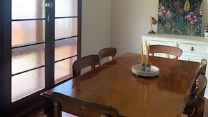 A dining area with a wooden table and chairs, a woven pendant light, a painting on the wall, and a frosted glass door.