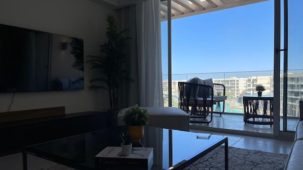 Un salon moderne doté d’un grand téléviseur, d’une table basse avec des livres, d’un canapé et d’un balcon aménagé avec du mobilier d’extérieur.