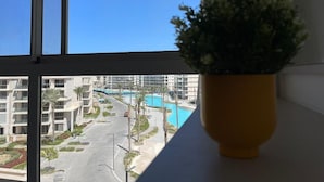 Une vue depuis une fenêtre sur un hôtel avec une piscine, des palmiers et un ciel bleu dégagé.