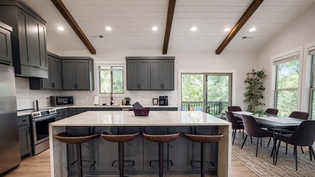 Een moderne keuken met donkere kastjes, een centraal kookeiland met barkrukken, een eethoek met tafel en stoelen, en grote ramen.